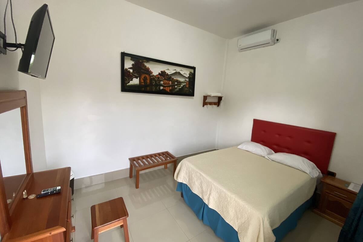 Ein Hotelzimmer mit Bett, Schreibtisch, Stuhl, kleinem Tisch, Wand-Fernseher, einem Gemälde und einer Klimaanlage.