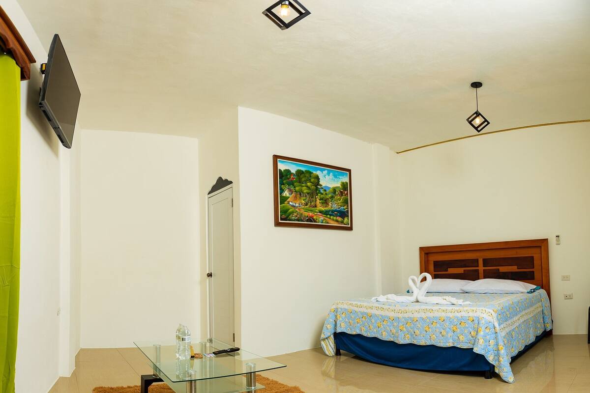 Ein Schlafzimmer mit einem Bett, einem Gemälde, einem Glastisch und einem an der Wand montierten Fernseher.