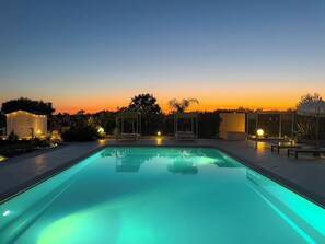Una piscina al anochecer con un atardecer de fondo, rodeada de mobiliario exterior y un pabellón.