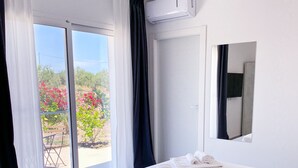 A hotel room with a bed, a window with curtains, an air conditioning unit, and a view of a garden with pink flowers. - Dimora Calauriia - Adults Only (Noto)