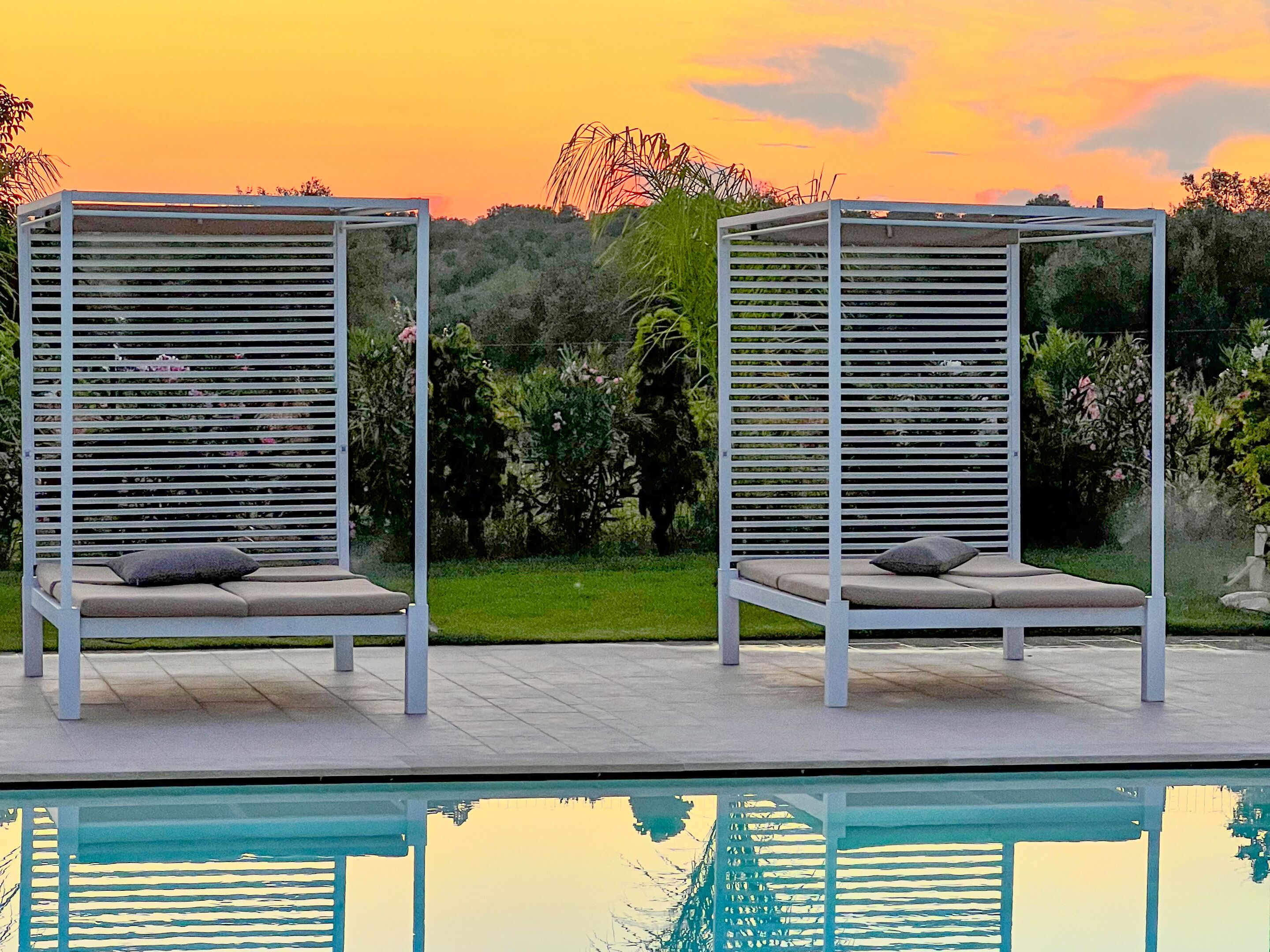 Zwei weiße Lounge-Sessel mit Kissen, aufgestellt am Pool mit Blick auf den umliegenden Garten und die Hügel im Hintergrund.