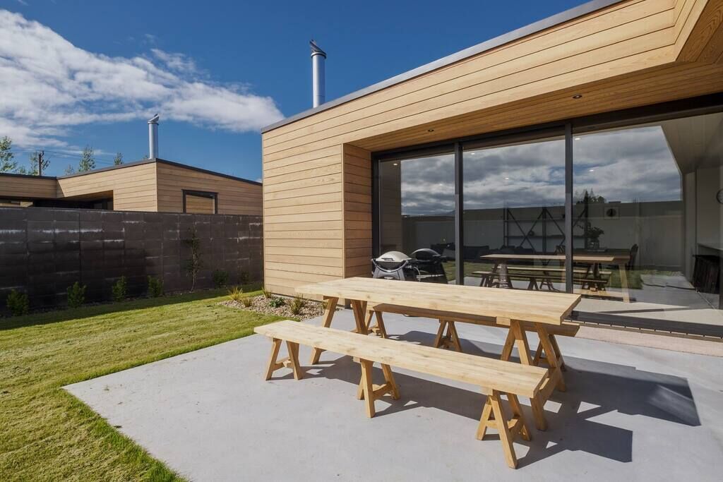 A modern house with a wooden exterior, a patio with a wooden table and benches, and a visible grill area.