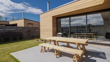 A modern house with a wooden exterior, a patio with a wooden table and benches, and a visible grill area.