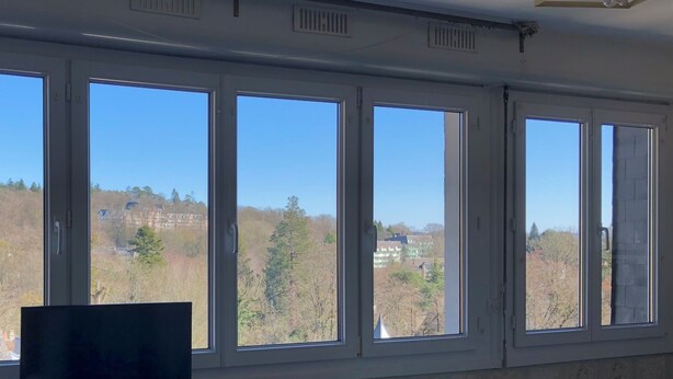 Una habitación con ventanas amplias que ofrecen vistas a una colina con árboles y un edificio con torre.