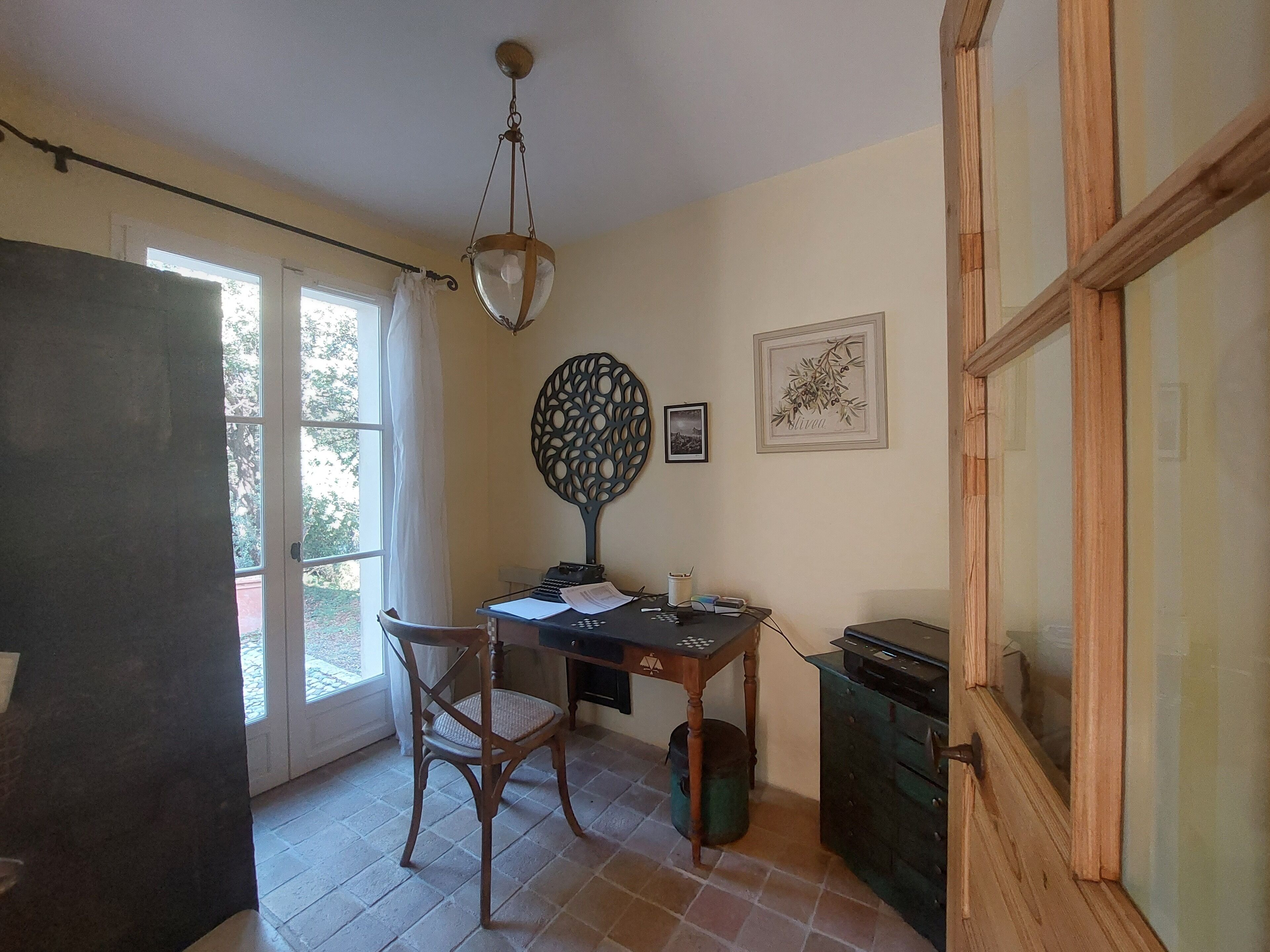 Una habitación con un escritorio de madera, una silla y una lámpara colgante. Se ve una ventana con cortinas, una puerta con paneles de vidrio y un cuadro en la pared.