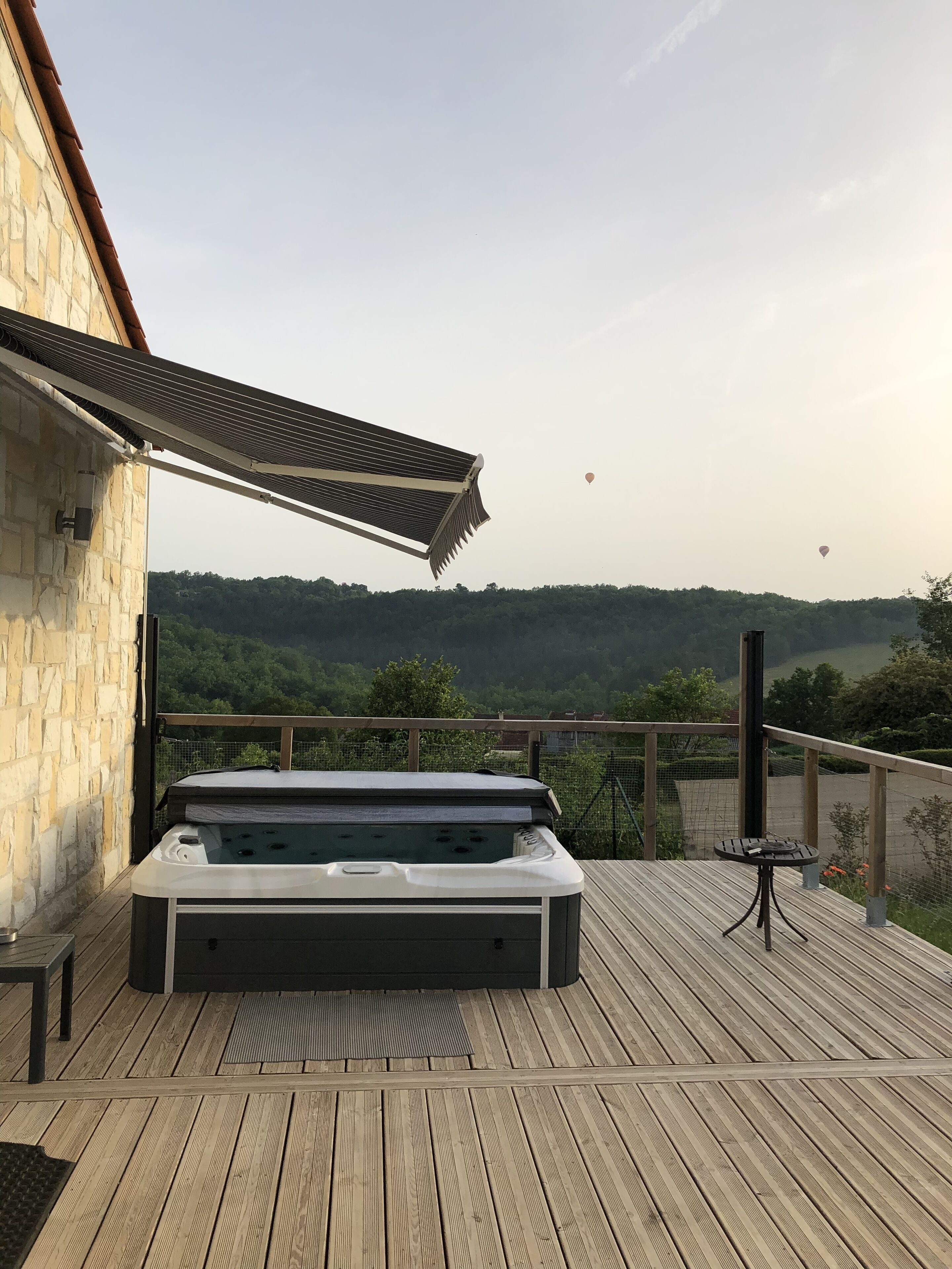 A hot tub on a wooden deck with a view of a forested hill and hot air balloons in the distance.