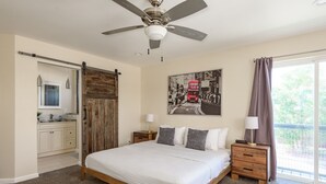 Una camera da letto con un letto grande, una ventola a soffitto, una finestra con tende, un comodino, un'armadio in legno e una zona cucina visibile tramite una porta scorrevole.