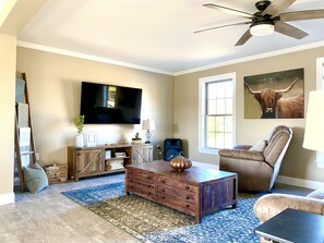 Un salon doté d’un téléviseur à écran plat, d’un meuble de rangement en bois, d’un tapis bleu et beige, d’un fauteuil en cuir et d’un ventilateur de plafond.