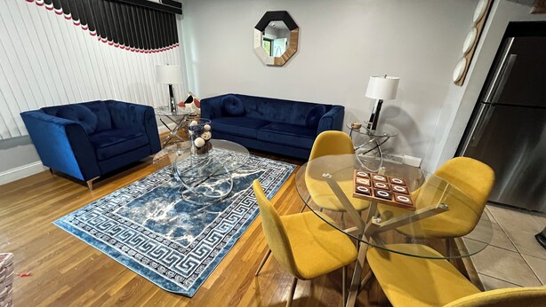 Un salon avec un canapé bleu, des fauteuils jaunes, une table basse en verre et un tapis bleu à motifs.