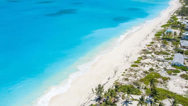 An aerial view of a beach with clear turquoise waters, white sand, and a row of houses with a dock extending into the water. - Colorful Beach Home w/ Ocean Front View + 2 Master Bedrooms (Exuma)