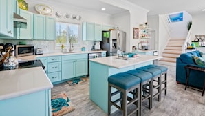 A modern kitchen with teal cabinetry, a central island, and a dining area with blue bar stools. - Colorful Beach Home w/ Ocean Front View + 2 Master Bedrooms (Exuma)