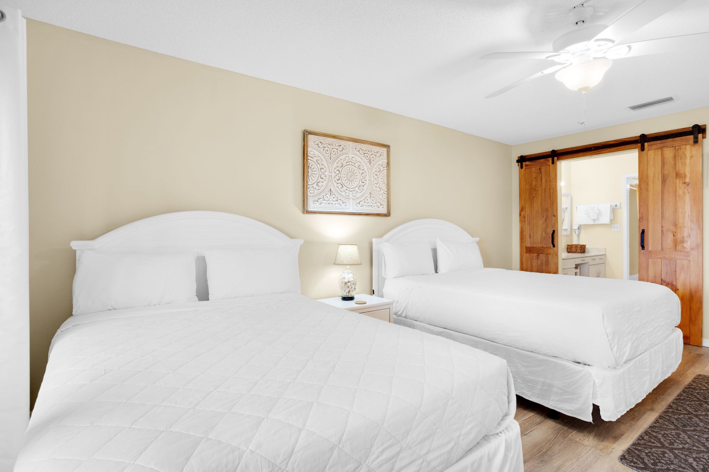 A hotel room with two beds, a ceiling fan, a bedside table with a lamp, and a wooden door.