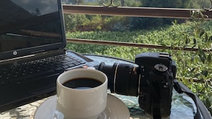 Un ordinateur portable, un appareil photo, une tasse de café et des lunettes de soleil sont posés sur une table, avec une vue sur les montagnes et la verdure.