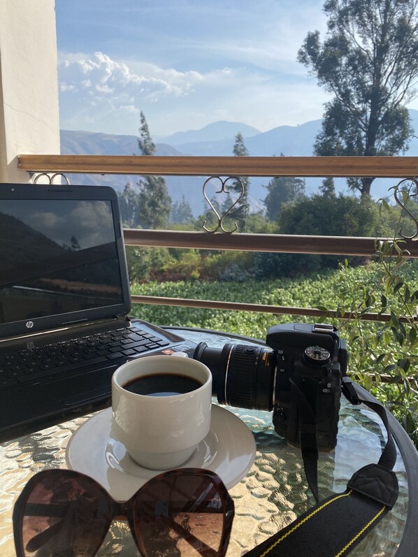 A laptop, a camera, a cup of coffee, and sunglasses on a table with a view of mountains and greenery.