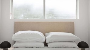 Una cama con cabecera color beige, ropa de cama blanca y dos mesitas de noche negras.