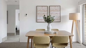 Un comedor con una mesa de madera, cuatro sillas, un jarrón con plantas, dos cuadros abstractos enmarcados, una lámpara de pie y una puerta que da a otra habitación.