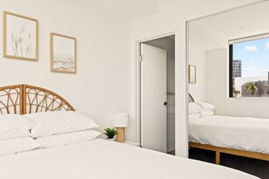 Une chambre à coucher avec un lit, une table de chevet, une petite plante et une fenêtre donnant sur des immeubles.