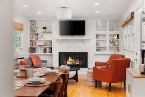 A modern living room with a fireplace, a flat-screen TV mounted above it, and a dining area with a table set for four.
