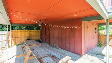 Un patio exterior con mesa y sillas de madera, una parrilla y un área cubierta con luces colgantes.