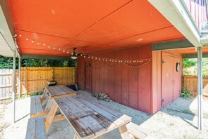 Un patio exterior con mesa y sillas de madera, una parrilla y un área cubierta con luces colgantes.