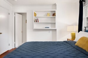 A bedroom with a large bed, a bedside table with a lamp, a bookshelf, and a door leading to another room.