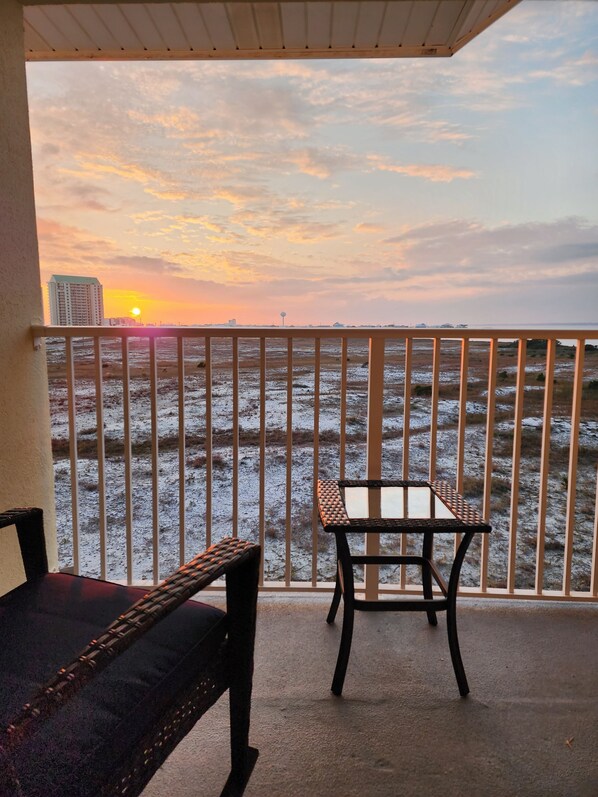 A balcony with a view of a sunset over a coastal landscape, featuring a table and a bench.