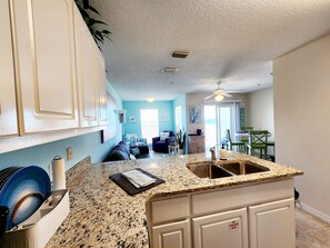 Una cocina con armarios blancos, encimera de granito y un lavabo. Hay un comedor con una mesa y sillas, y una sala de estar con un sofá y una mesita de centro.