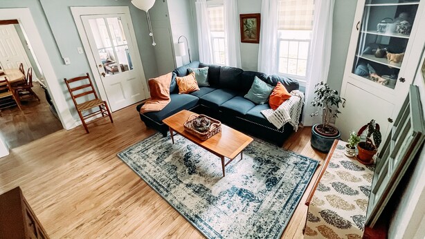 Un salon comprenant un canapé d’angle, une table basse, un coin repas avec des chaises, un ventilateur de plafond et une fenêtre avec des rideaux.
