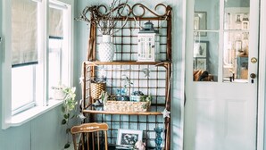 Une pièce avec un plafond en bois, une chaise en osier, une étagère en osier contenant divers objets et une porte dotée d’un miroir.