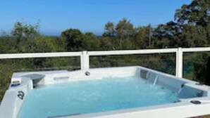 Un jacuzzi con revestimiento blanco, rodeado por una terraza de madera y una barandilla blanca, con árboles y un cielo despejado de fondo.