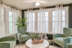 A room with a ceiling fan, large windows with sheer curtains, and green upholstered chairs around a round table with a woven basket and potted plants. - The Vedder Cottage (St. Augustine)