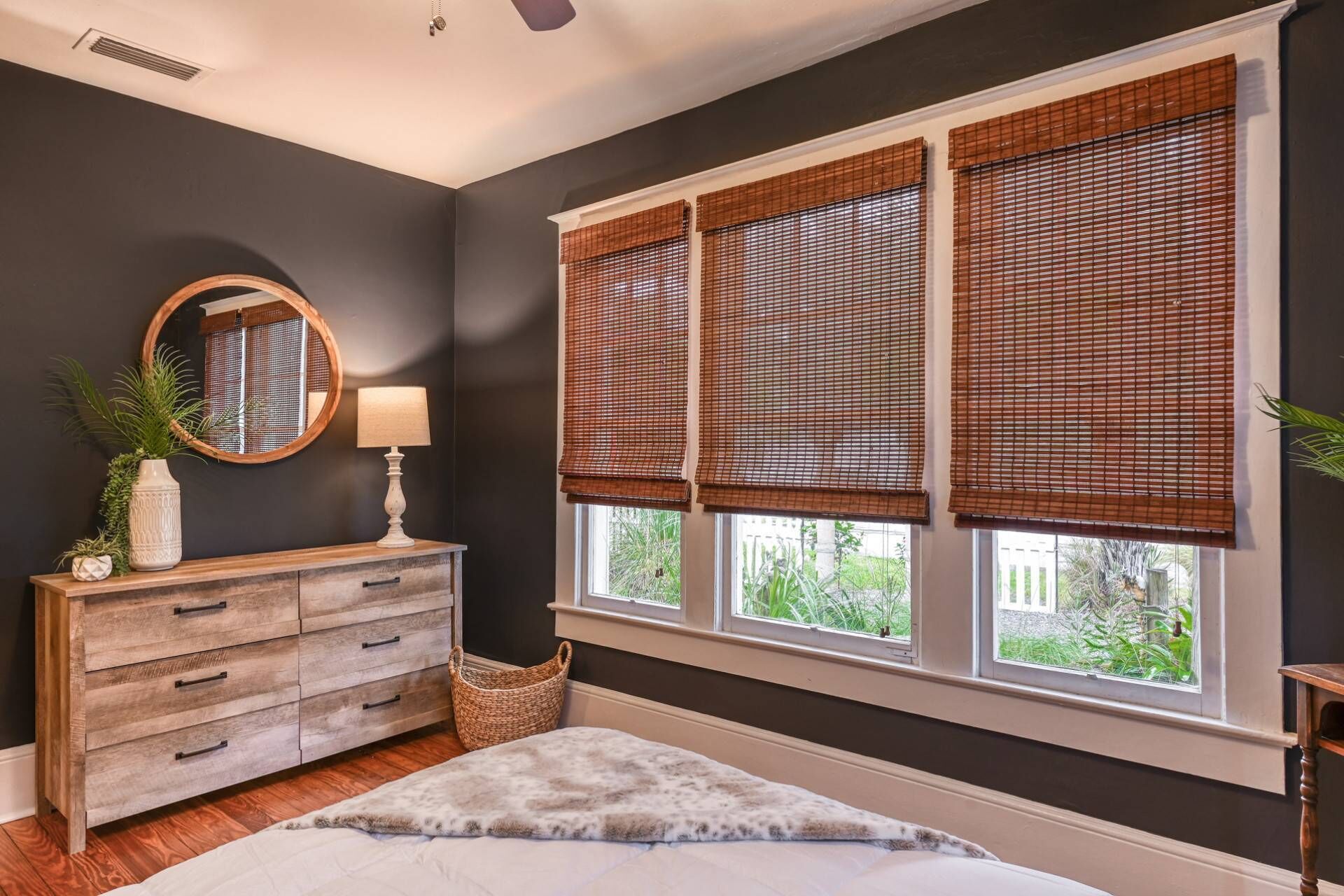 Quarto com uma grande janela, cómoda de madeira, espelho e mesa de cabeceira.