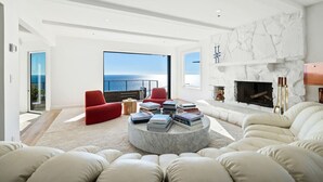 Un salon moderne avec une cheminée, une table basse ronde sur laquelle sont posés des livres, des canapés rouges et blancs, et une vue sur l’océan.