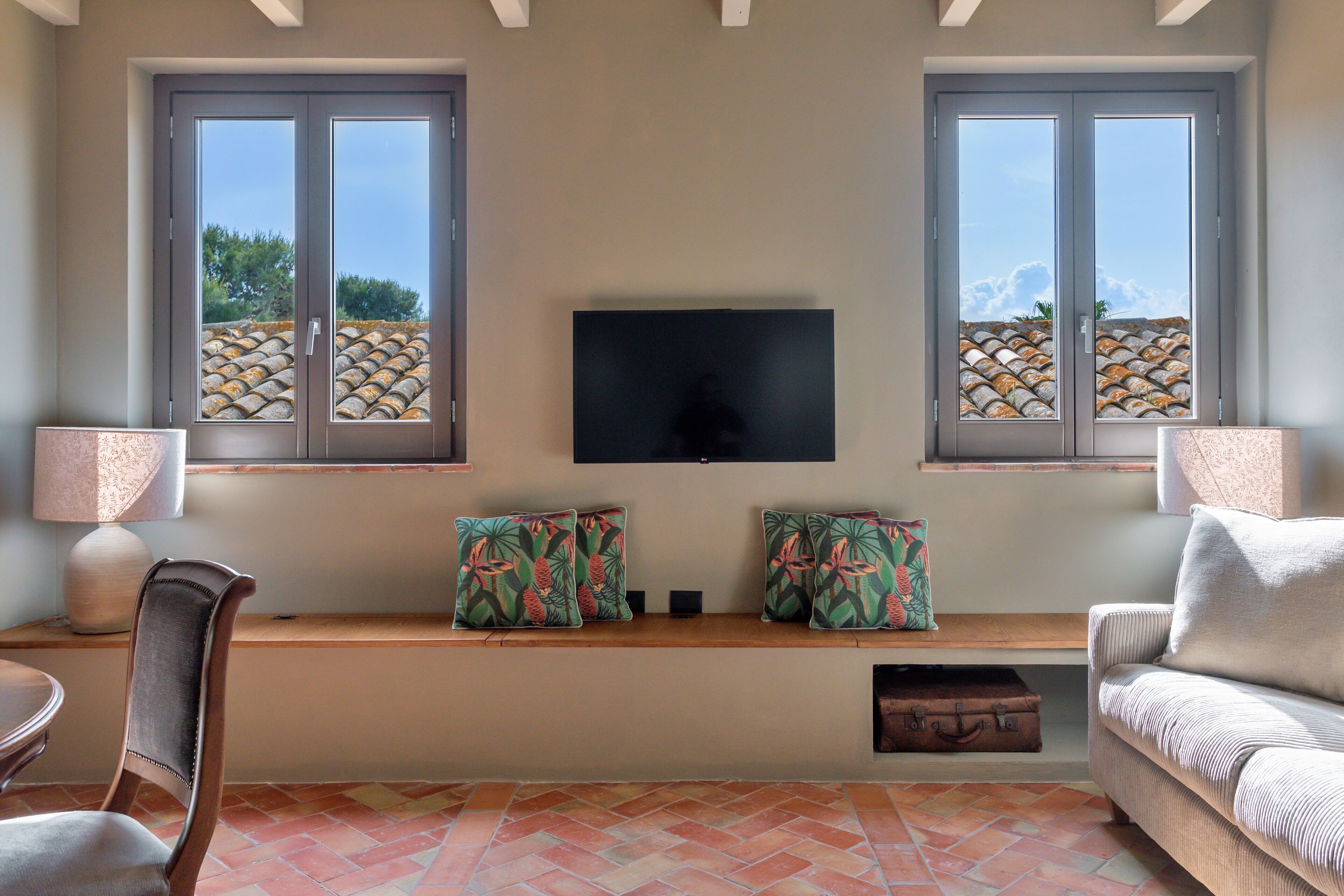 A living room with a television on the wall, two windows with a stone wall view, a bench with cushions, a sofa, and a chair.