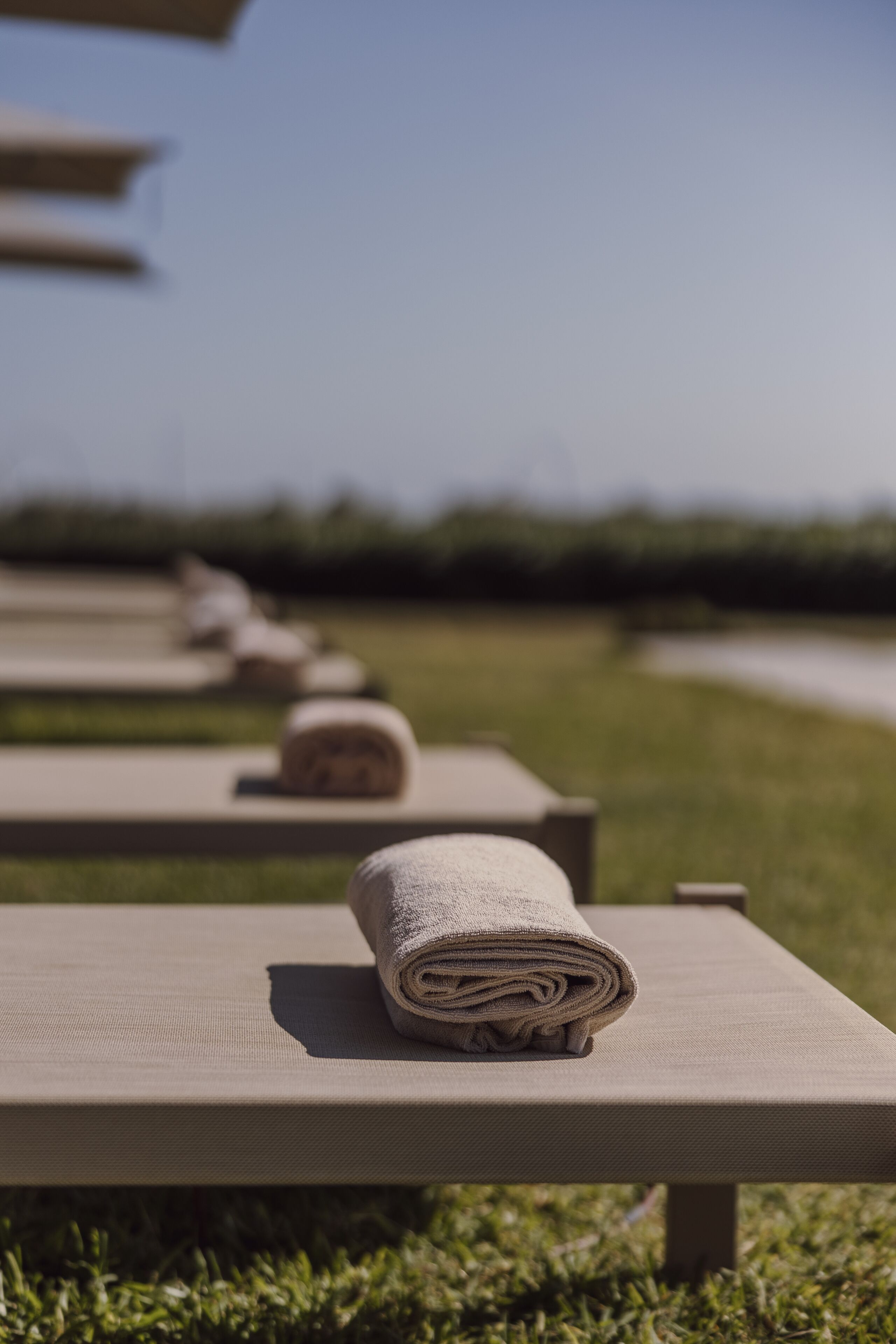 A row of outdoor lounge chairs with towels, set against a grassy area with a clear sky.