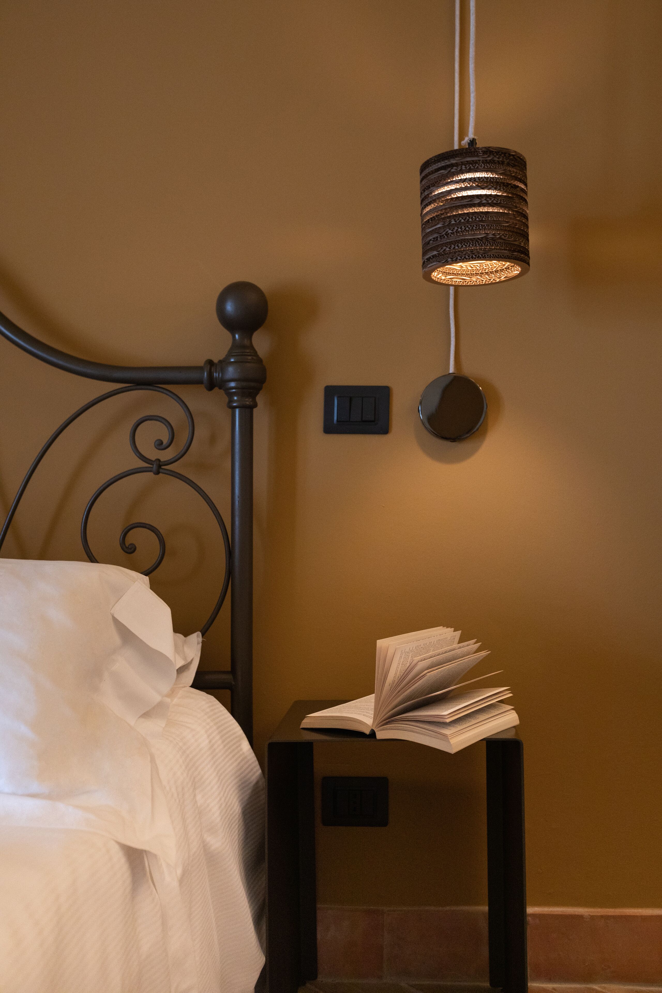 A bed with a white comforter, a bedside table with an open book, and a wall-mounted lamp.