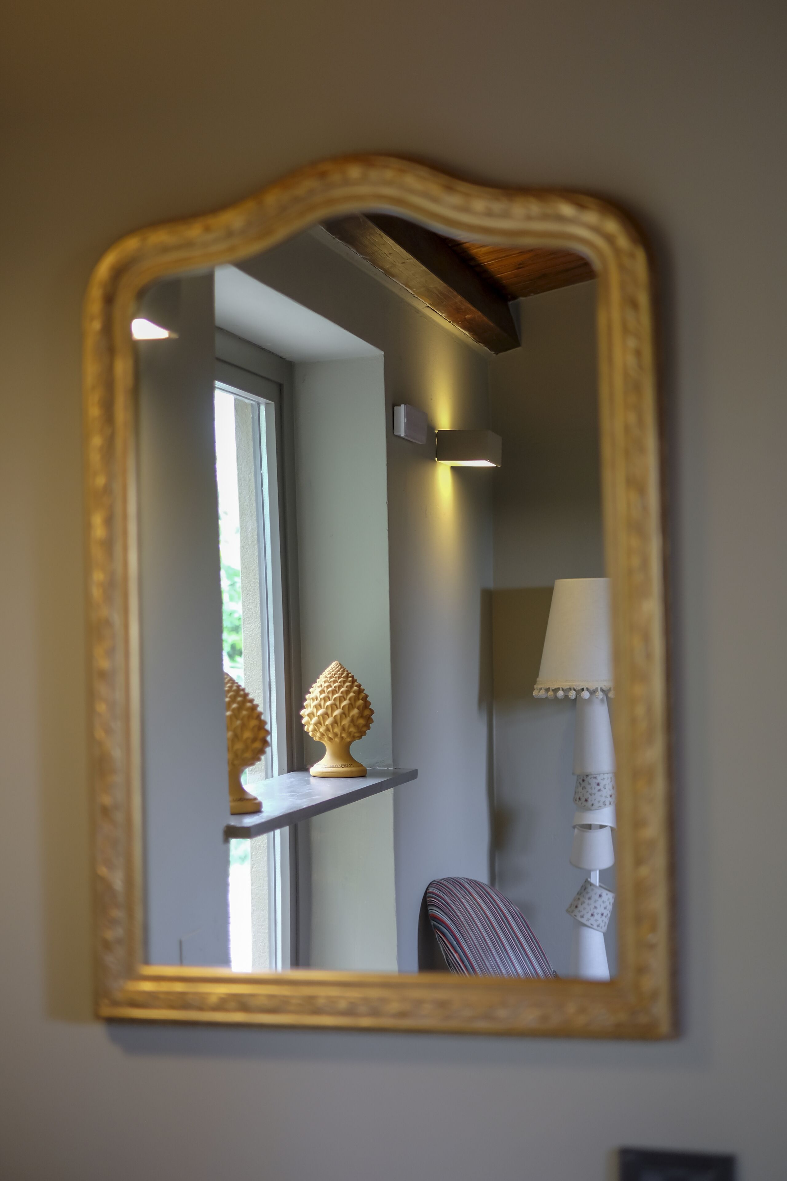 A mirror with a gold frame reflecting a room with a lamp, a chair, and a shelf with a decorative item.