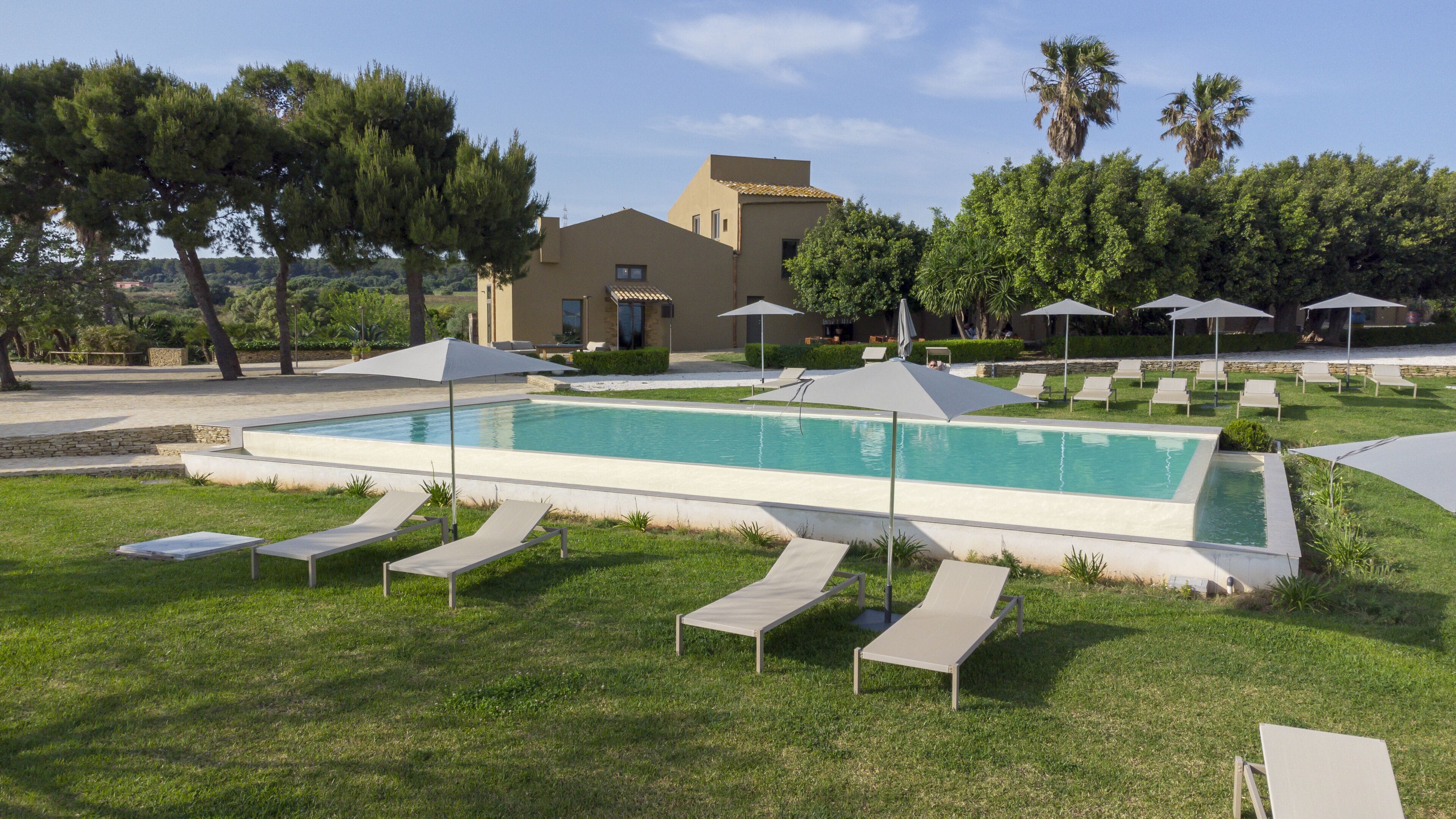 Una piscina con sillas de descanso y sombrillas, rodeada de follaje y un edificio al fondo.