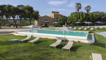 Una piscina con sillas de descanso y sombrillas, rodeada de follaje y un edificio al fondo.