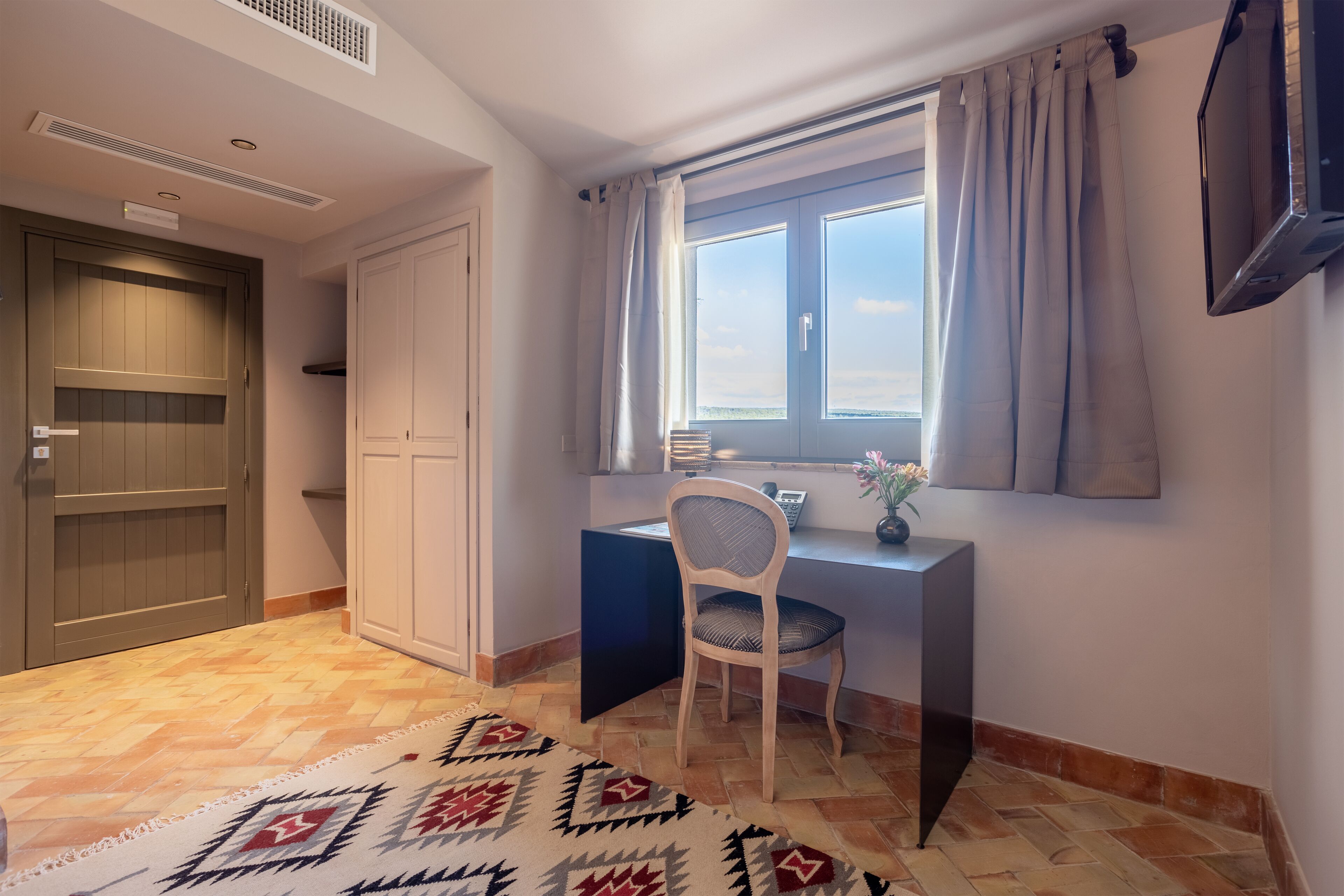 A room with a desk, chair, and a view through a window with curtains.