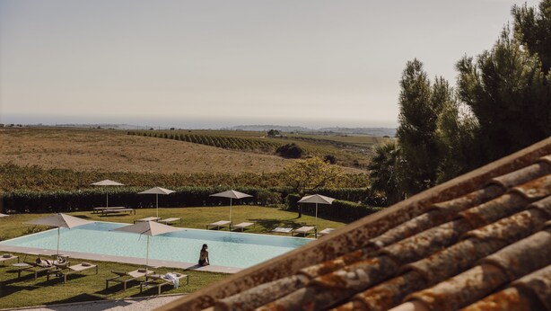 Zona de piscina con sillas de descanso y sombrillas, rodeada de un jardín ajardinado y vistas a colinas distantes.