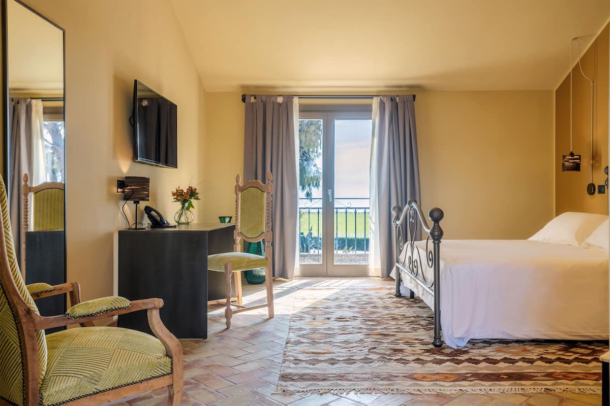 A bedroom with a bed, a desk with a chair, a mirror, and a view of the outdoors through a sliding door.