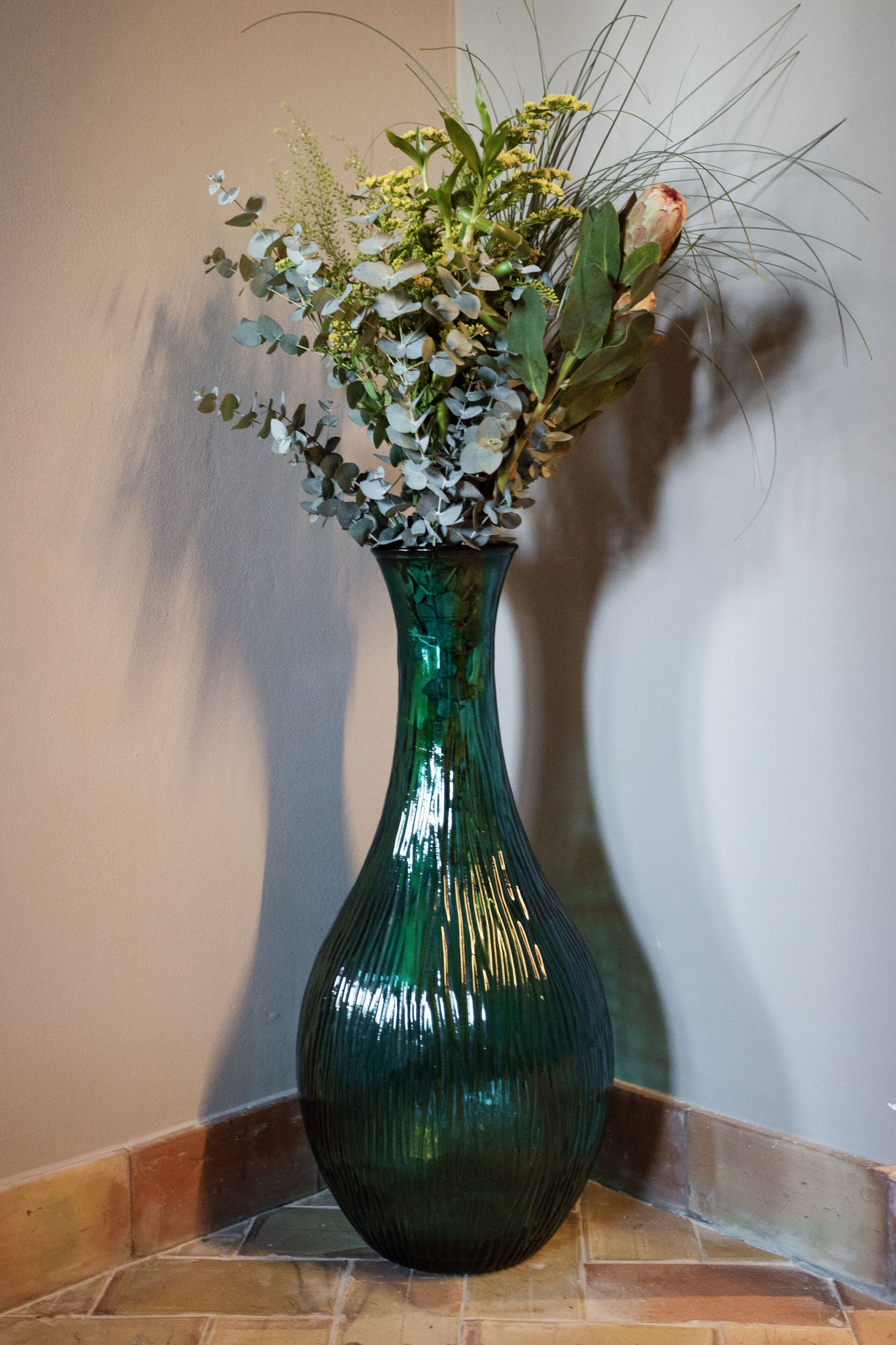 A green vase with a floral arrangement on a tiled floor against a wall.
