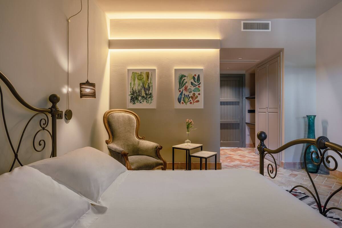 A bedroom with a bed, a chair, a small table with a vase, and two wall-mounted artworks.