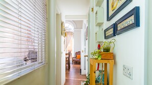 Un couloir avec un plancher en bois, une fenêtre avec des stores, une porte et un mur orné de tableaux et sur lequel est posée une petite table avec une plante.