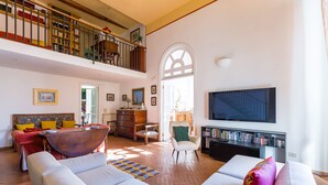Un salon spacieux avec un haut plafond, un balcon agrémenté d’étagères à livres, une grande télévision et un coin salon confortable.