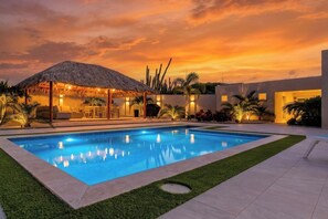 A swimming pool with a thatched-roof structure, surrounded by a well-maintained lawn and palm trees.