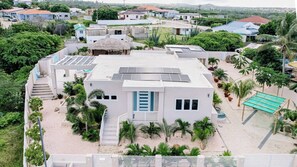 A residential area with a house featuring a solar panel roof, a thatched-roof structure, and a sandy courtyard with palm trees. - Incredible 4BR w/ Private Pool near Palm Beach (Noord)