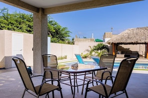 A covered outdoor seating area with a pool, lounge chairs, and a thatched-roof structure.