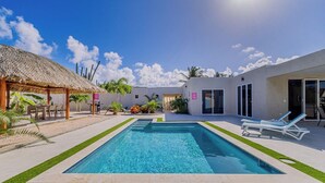 Une piscine dotée d’une structure en chaume et de chaises longues, entourée de verdure et surplombée par un ciel dégagé.